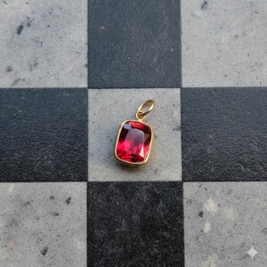 May include: A gold pendant with a rectangular, deep red gemstone. The gemstone is set in a gold bezel and has a small gold loop for a chain. The pendant is displayed on a black and gray checkered surface.