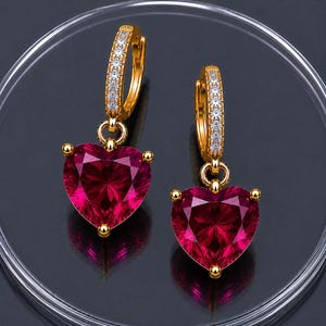 May include: A pair of gold hoop earrings featuring a row of small clear stones along the front of the hoop and a large, faceted, heart-shaped deep red gemstone dangling from the base of each earring, secured by a small gold metal prong setting.