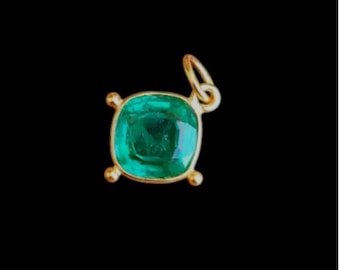 May include: A gold pendant with a square, emerald-cut green gemstone. The pendant has a small loop at the top for a chain and four small gold accents at the corners of the gemstone. The background is black.