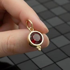 Puede incluir: Un colgante de oro con una gema redonda de color rojo intenso. El colgante tiene un diseño sencillo y elegante con un pequeño lazo para una cadena y un grupo de tres pequeñas cuentas de oro en la parte inferior.