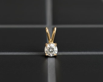 May include: A gold pendant with a single, round, brilliant-cut diamond. The diamond is set in a four-prong setting, and the pendant has a simple, elegant design. The pendant is displayed on a black surface.
