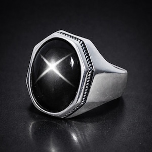 May include: A silver ring featuring a large, oval black gemstone with a star-shaped pattern. The ring has a thick band and a decorative border. The gemstone's surface reflects light, creating a striking visual effect.