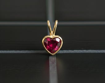 Op de afbeelding: Een hartvormige hanger van goudkleurig metaal met een helderrode edelsteen. De hanger heeft een eenvoudig, elegant ontwerp met een gepolijste afwerking. De gefacetteerde edelsteen vangt het licht op en creëert een sprankelend effect. Voor een ketting.