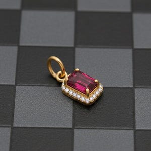 May include: A gold pendant with a rectangular, deep red gemstone at its center, surrounded by a halo of small, clear gemstones. The pendant has a loop at the top for a chain. The background is a checkered pattern of black and gray squares.
