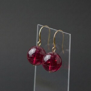 May include: A pair of gold-colored drop earrings featuring large, round, deep red beads. The beads have a translucent quality, catching the light. The earrings are displayed against a neutral background.
