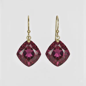 May include: A pair of gold-toned drop earrings featuring large, square-cut, deep red gemstones. The gemstones have multiple facets that catch the light. The earrings have a simple hook design.