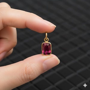 May include: A small, rectangular pendant with a deep pink gemstone set in a gold-colored frame. The pendant is held between two fingers against a dark, textured background. The gemstone has a rectangular cut.