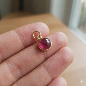 May include: A small, round pendant with a vibrant, deep pink gemstone set in a gold-colored metal. The pendant has a simple, elegant design with a loop at the top for attaching to a chain. The gemstone is approximately 0.3 inches in diameter.