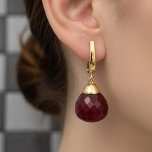 May include: A close-up of a gold earring with a teardrop-shaped, faceted, deep red gemstone. The earring is attached to the ear with a gold clasp. The gemstone is a rich, dark red color, and the gold has a polished finish.
