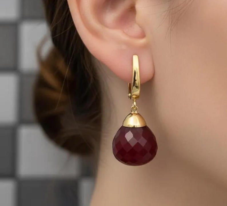 May include: A close-up of a gold earring with a teardrop-shaped, faceted, deep red gemstone. The earring is attached to the ear with a gold clasp. The gemstone is a rich, dark red color, and the gold has a polished finish.