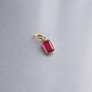May include: A gold pendant with a rectangular, deep red gemstone. The gemstone is set in a gold bezel and has a polished surface. The pendant has a small loop for attaching to a chain or bracelet.