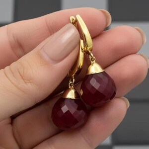 May include: A pair of gold-colored drop earrings with faceted, deep red teardrop-shaped gemstones. The earrings have a lever back closure and a gold cap at the top of each gemstone. The earrings are held up by a hand.