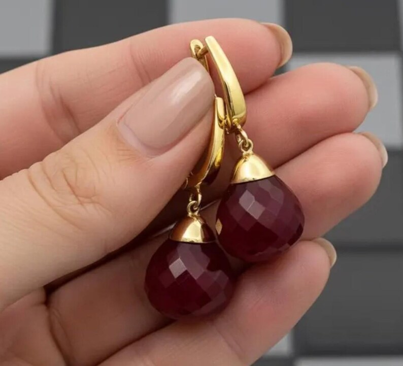 May include: A pair of gold-colored drop earrings with faceted, deep red teardrop-shaped gemstones. The earrings have a lever back closure and a gold cap at the top of each gemstone. The earrings are held up by a hand.