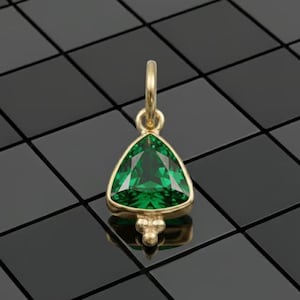 Könnte beinhalten: Ein goldener Anhänger mit einem dreieckigen, facettierten Smaragd-Edelstein. Der Smaragd hat eine leuchtend grüne Farbe und ist in eine goldene Lünette gefasst. Der Anhänger hat eine kleine Schlaufe für eine Kette und drei kleine Goldkügelchen.