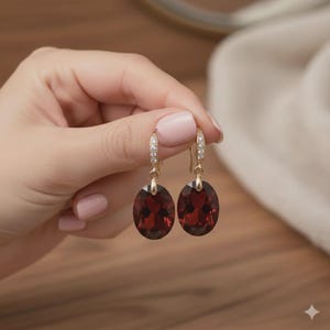 Puede incluir: Un par de pendientes colgantes dorados. Cada pendiente presenta una gran piedra preciosa ovalada de color rojo oscuro suspendida de una sección más pequeña y curvada adornada con pequeños cristales transparentes. Los pendientes son sostenidos por una mano.