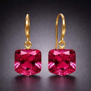 May include: A pair of gold-colored drop earrings featuring large, faceted, square-cut, ruby-red gemstones. The earrings have a simple, elegant design with a hook closure. The gemstones reflect light, creating a sparkling effect against a dark background.