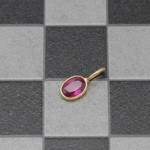 May include: A gold pendant featuring a large, oval, magenta gemstone. The pendant has a simple, elegant design with a smooth, polished gold setting. The gemstone is the focal point, reflecting a vibrant hue. The pendant is set against a checkered background.