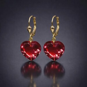 May include: A pair of heart-shaped earrings with a faceted, deep red gemstone. The earrings have a gold-colored leverback closure. The heart-shaped gems reflect light, creating a sparkling effect. The earrings are displayed against a dark background.