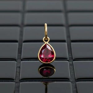 May include: A teardrop-shaped pendant with a faceted, deep red gemstone set in a gold-colored bezel. The pendant hangs from a small, gold-colored loop. The background is a glossy, black tiled surface.