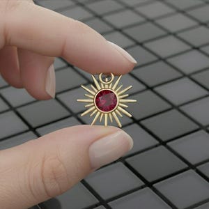 Puede incluir: Un colgante dorado con forma de sol, con una gema roja redonda en el centro. El colgante se sostiene entre los dedos sobre un fondo oscuro y embaldosado. El diseño de sol tiene rayos puntiagudos que se extienden hacia afuera.