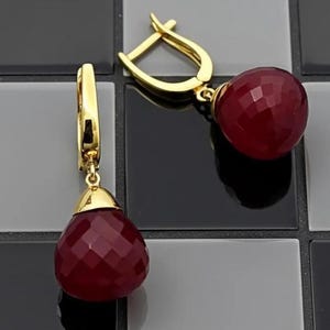 May include: A pair of gold-colored earrings with a drop design. Each earring features a faceted, deep red, teardrop-shaped gemstone. The earrings have a lever-back closure and a gold-colored cap that connects the gemstone to the earring.