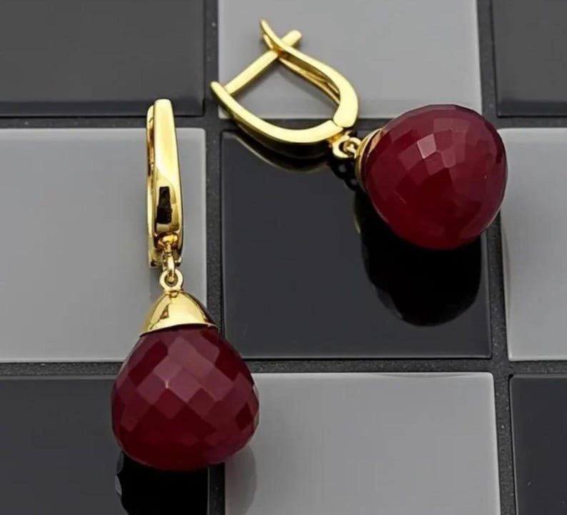 May include: A pair of gold-colored earrings with a drop design. Each earring features a faceted, deep red, teardrop-shaped gemstone. The earrings have a lever-back closure and a gold-colored cap that connects the gemstone to the earring.