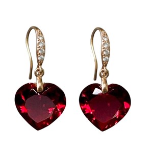 Pode incluir: Um par de brincos pendurados dourados. Cada brinco apresenta um pendente de cristal vermelho escuro em forma de coração. Os ganchos dos brincos são dourados e têm uma pequena fileira de cristais transparentes.