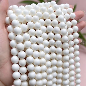 May include: A collection of smooth, opaque white spherical beads arranged in multiple vertical strands. The beads have a uniform, glossy finish and are strung together. The text EllaJewelryStarGifts is visible on the right side.