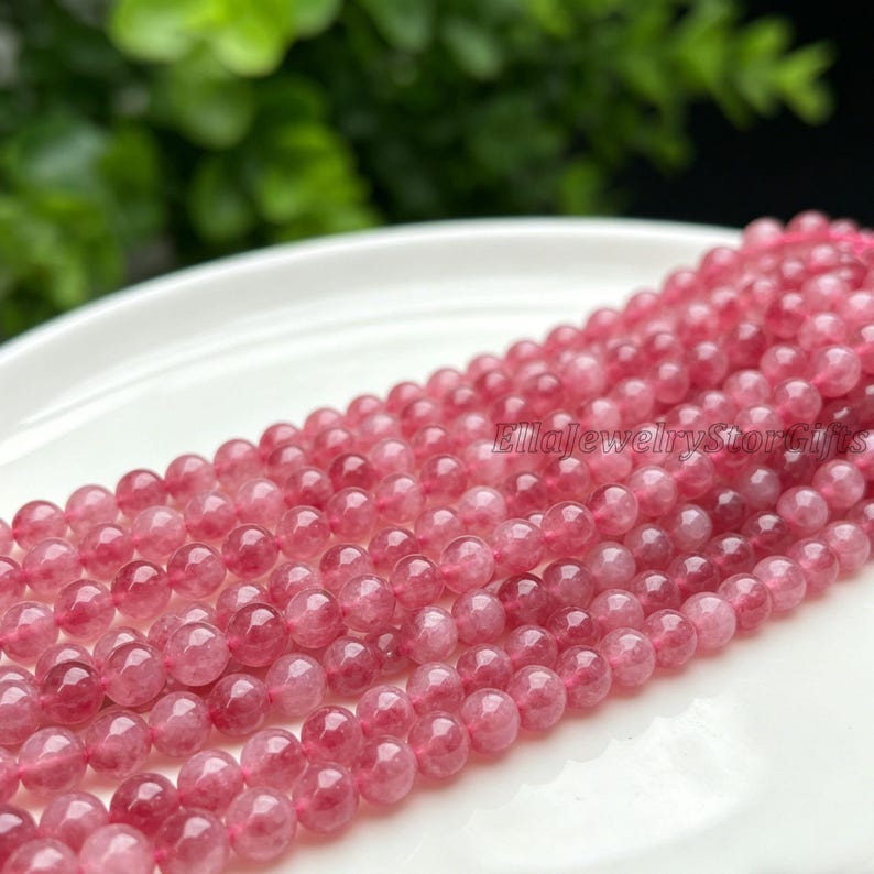Puede incluir: Primer plano de un hilo de cuentas de piedras preciosas rosas. Las cuentas son redondas y tienen una apariencia transl&uacute;cida. El texto "EllajewelryStoreGifts" es visible. Las cuentas est&aacute;n dispuestas sobre una superficie blanca.