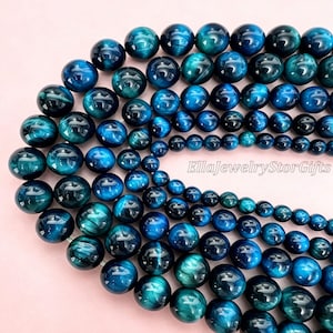 Puede incluir: Una colección de cuentas de piedra preciosa ojo de tigre azul, pulidas y esféricas, dispuestas en un patrón curvo. Las cuentas presentan un acabado brillante con tonos azul marino profundo y turquesa. El texto EllaJewelryStorGifts es visible.