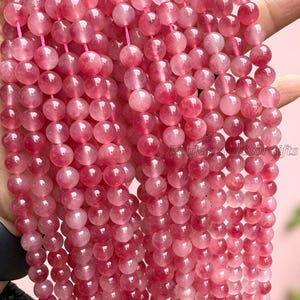 Puede incluir: Cadenas de cuentas de piedras preciosas rosas. Cada cuenta redonda tiene una apariencia transl&uacute;cida con inclusiones de color rosa oscuro. Las cuentas est&aacute;n ensartadas en un cord&oacute;n rosa. Las cuentas se sostienen en una mano.