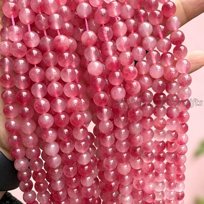 Puede incluir: Cadenas de cuentas de piedras preciosas rosas. Cada cuenta redonda tiene una apariencia transl&uacute;cida con inclusiones de color rosa oscuro. Las cuentas est&aacute;n ensartadas en un cord&oacute;n rosa. Las cuentas se sostienen en una mano.