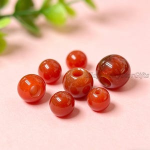 Puede incluir: Una colección de cuentas redondas de piedra preciosa rojo-naranja pulidas. Algunas cuentas tienen un agujero en el centro. Las cuentas varían ligeramente en tamaño y color, con un acabado brillante. Las cuentas están dispuestas sobre una superficie rosa.