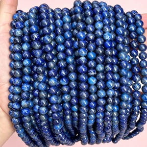 Könnte beinhalten: Eine Sammlung mehrerer Stränge aus runden, polierten Lapislazuli-Perlen. Die Perlen haben eine tief königsblaue Farbe mit natürlichen dunkelgrauen und schwarzen Einschlüssen und sind in vertikalen Reihen angeordnet, um ihre glatte Textur zu zeigen.