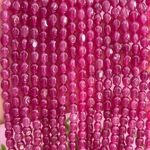 Puede incluir: Múltiples hilos verticales de cuentas lisas, pulidas y de forma ovalada en un vibrante color rosa magenta. Cada cuenta presenta un acabado brillante y translúcido que refleja la luz, creando una apariencia uniforme en todo el conjunto.