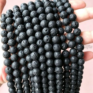 May include: Close-up of black lava rock beads. The beads are spherical with a textured, porous surface. The dark charcoal beads are held in a hand and appear to be strung together, ready for crafting.