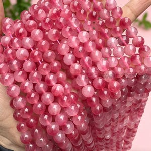 Puede incluir: Primer plano de hileras de cuentas de cuarzo rosa. Las cuentas redondas y transl&uacute;cidas presentan tonos rosados claros y oscuros. Est&aacute;n ensartadas en un hilo rosa. Las cuentas son lisas y pulidas, reflejando la luz. La imagen destaca el color y la textura.