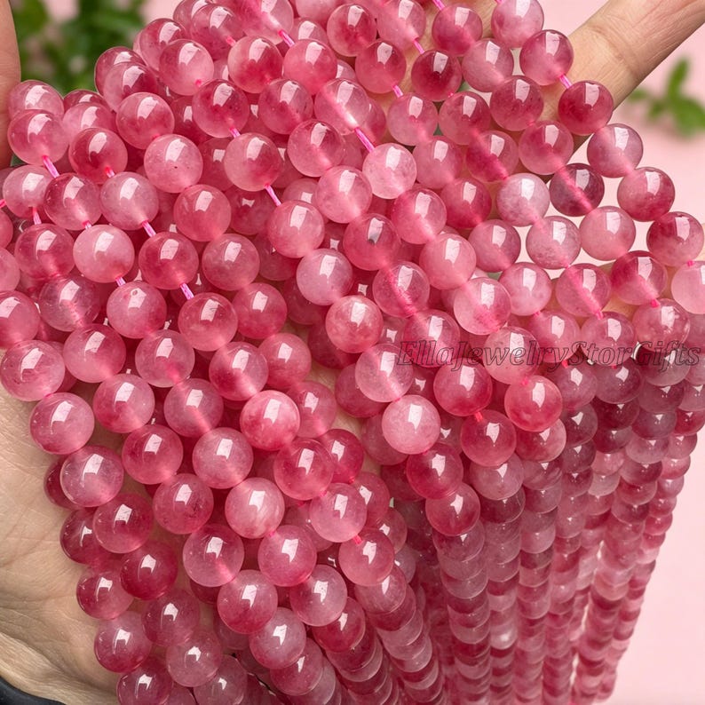 Puede incluir: Primer plano de hileras de cuentas de cuarzo rosa. Las cuentas redondas y transl&uacute;cidas presentan tonos rosados claros y oscuros. Est&aacute;n ensartadas en un hilo rosa. Las cuentas son lisas y pulidas, reflejando la luz. La imagen destaca el color y la textura.