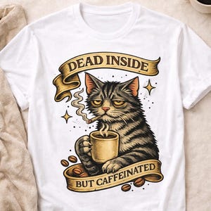 May include: White t-shirt featuring a grumpy cat illustration. The cat, holding a coffee mug and smoking, is surrounded by coffee beans and a banner that reads "DEAD INSIDE BUT CAFFEINATED".