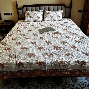 Può includere: Un letto con una trapunta bianca e federe abbinate con un motivo a cammello ripetuto in marrone e arancione. Il letto ha una testiera in legno scuro. Un laptop e un telefono sono sulla trapunta.