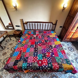 May include: A patchwork quilt in a bedroom setting. The quilt features a vibrant mix of colors and patterns, including red, blue, purple, and yellow. The bed has a wooden frame and a padded headboard. The room has a neutral wall color and decorative lighting.