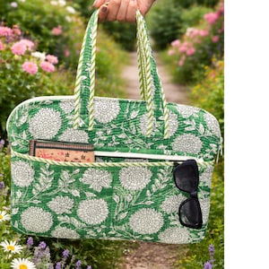 Può includere: Borsa tote con stampa floreale verde con dettagli bianchi. La borsa presenta un grande motivo floreale, una tasca frontale e una chiusura con cerniera. Sono visibili occhiali da sole neri e un piccolo portafoglio.