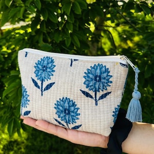May include: A quilted zippered pouch in an off-white fabric featuring a repeating blue floral print. The pouch includes a white zipper closure with a matching blue fabric tassel attached to the pull, showcasing a textured, stitched surface.