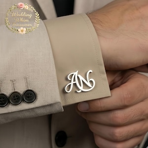 May include: A pair of silver-toned metal cufflinks featuring the intertwined letters AN in a cursive script font. The cufflinks are attached to the beige fabric cuff of a long-sleeved shirt, which is worn under a light beige textured blazer.