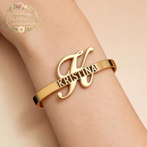 Può includere: Un bracciale rigido in metallo color oro caratterizzato da una grande lettera K corsiva stilizzata integrata nella fascia. Il nome KRISTINA è ritagliato in lettere maiuscole al centro della barra orizzontale che si estende dal design della K.