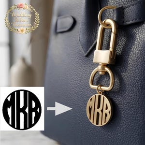 Może przedstawiać: Złoty brelok do torby z monogramem z inicjałami "MRR" w okrągłym wzorze. Brelok jest przymocowany do złotego zapięcia, wiszącego na granatowej torebce. Obraz zawiera również logo z tekstem "Wedding Whim Treasures".