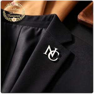 Könnte beinhalten: Nahaufnahme eines schwarzen Blazers mit einer silberfarbenen Anstecknadel. Die Anstecknadel zeigt die verschlungenen Buchstaben "NC". Der Blazer ist aus einem glatten Stoff gefertigt und Teil eines Anzugs. Das Bild ist gut beleuchtet und die Details sind scharf.