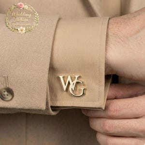 Op de afbeelding: Een goudkleurige manchetknop met de in elkaar grijpende letters WG, bevestigd aan de manchet van een beige overhemd met lange mouwen. Het overhemd wordt gedragen onder een beige blazer met een zichtbare knoop. Een bloemrijk logo met de tekst Wedding Whim Treasures is aanwezig.