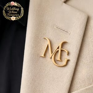 May include: A gold-toned metal brooch featuring the interlocking letters M and G in a serif font, pinned to a textured beige fabric lapel. The upper left corner displays a circular logo with floral accents and the text Wedding Whim Treasures.