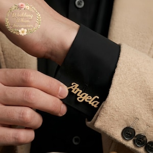 Op de afbeelding: Een goudkleurige manchetknop met de naam Angela in een cursief lettertype, bevestigd aan de zwarte stoffen manchet van een overhemdmouw, gedragen onder een beige gestructureerde blazer met donkere knopen.